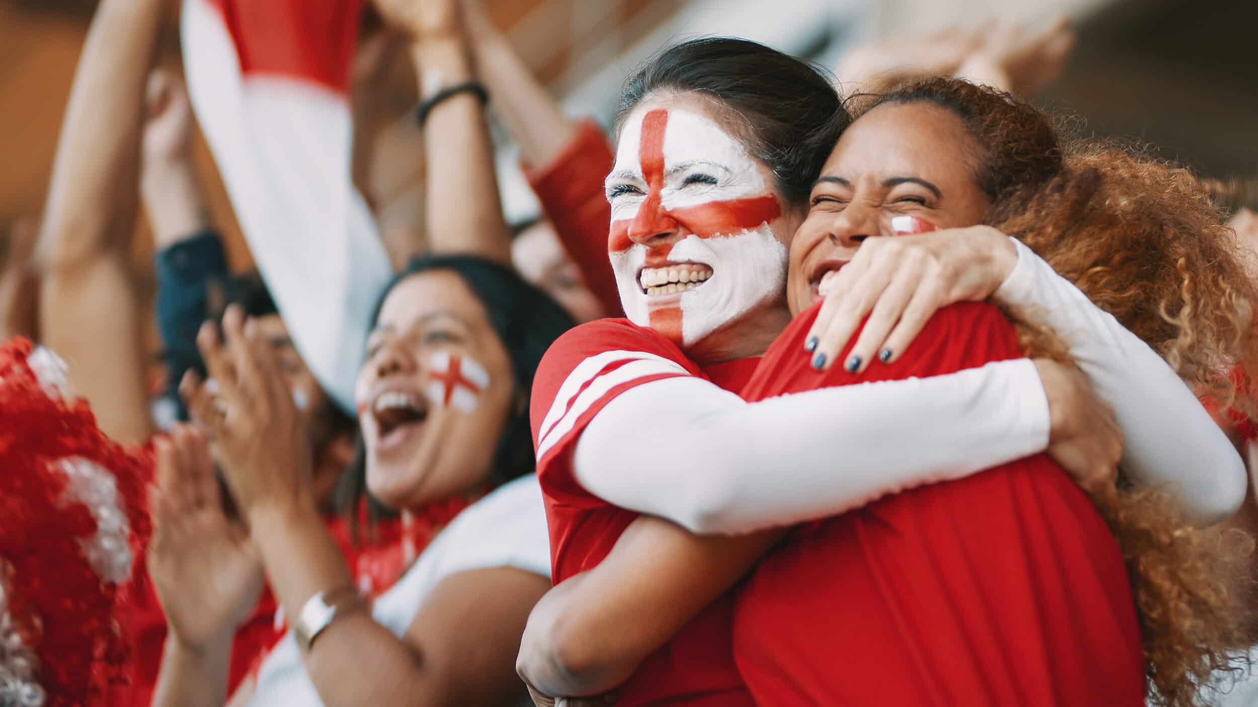 Two women in red shirts with England flag facepaint on their faces hugging each other and smiling showcasing hospitality technology solutions for stadiums.